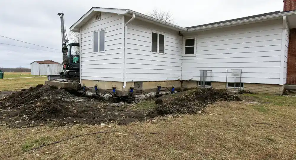 Foundation repair crew installing push piers in excavation trenches along a 1960s brick ranch home, with hydraulic equipment and reddish-brown Midwest clay soil visible
