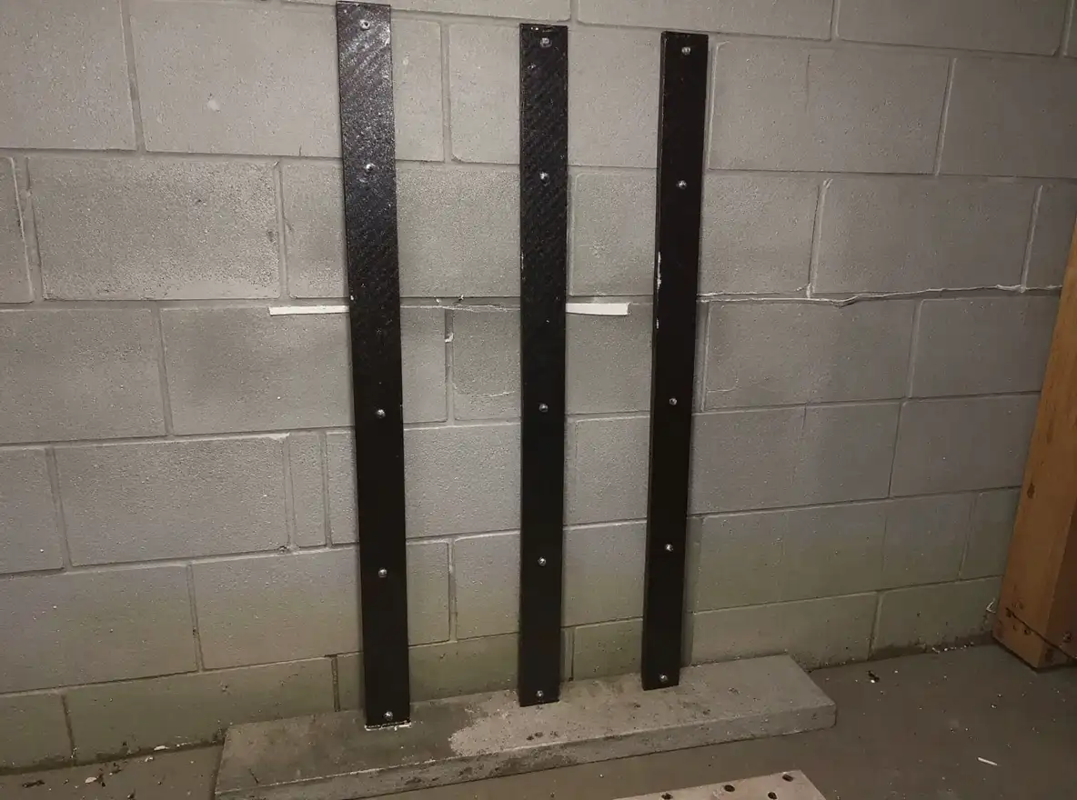 Three parallel carbon fiber straps bonded vertically to a concrete block basement wall with epoxy adhesive, bridging a horizontal crack to prevent further bowing