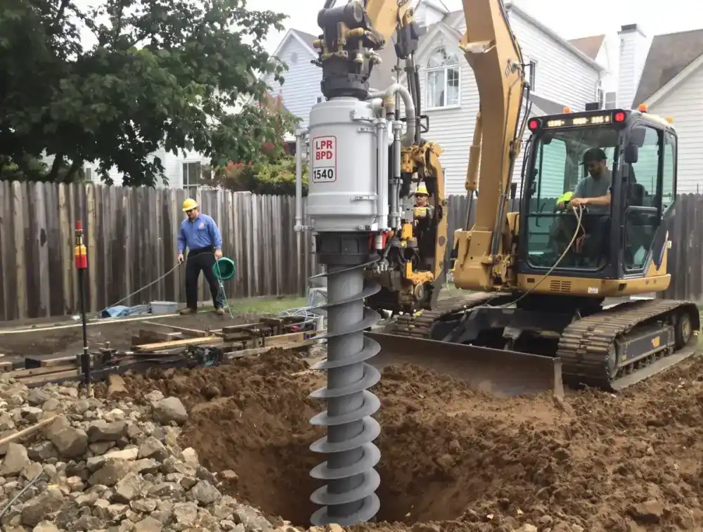 Helical pier shaft with spiral helix plates being screwed into clay soil by hydraulic torque equipment at a residential foundation, demonstrating the rotation-based installation method used for lighter structures and restricted-access sites