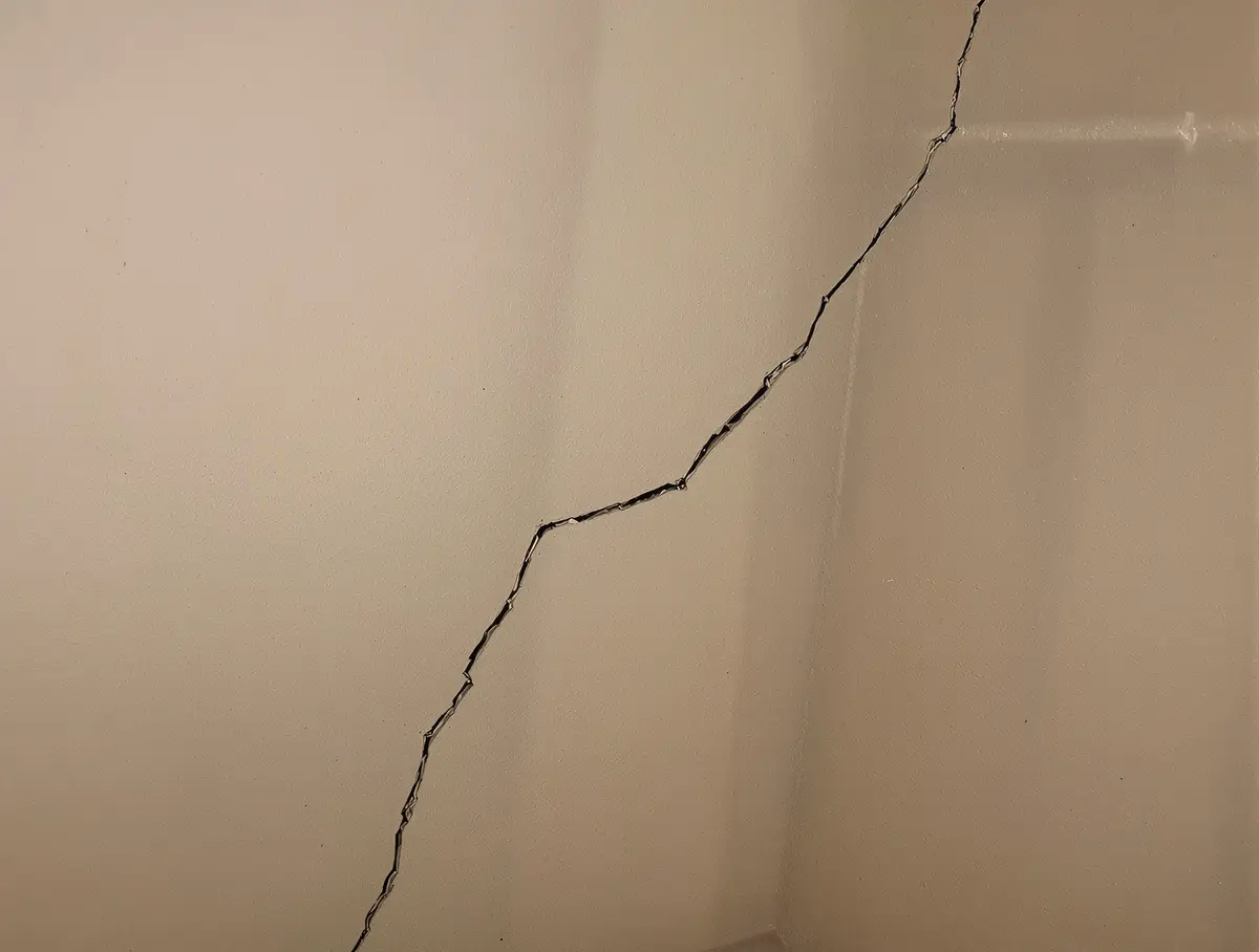 Diagonal crack extending at 45 degrees from a basement window corner in a poured concrete foundation wall, widening as it radiates outward to indicate differential settlement where one section of the foundation has dropped relative to another